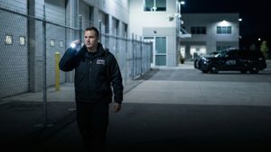 Apex Security Mitarbeiter führt nachts einen Streifendienst auf einem umzäunten Firmengelände durch und leuchtet mit einer Taschenlampe den Bereich ab, im Hintergrund stehen Einsatzfahrzeuge.
