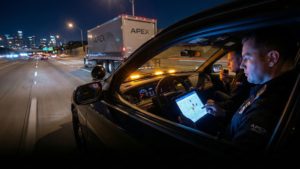 Apex Security Mitarbeiter begleiten nachts einen Lkw auf der Autobahn, überwachen den Transport per Tablet mit GPS-Karte aus dem Begleitfahrzeug.